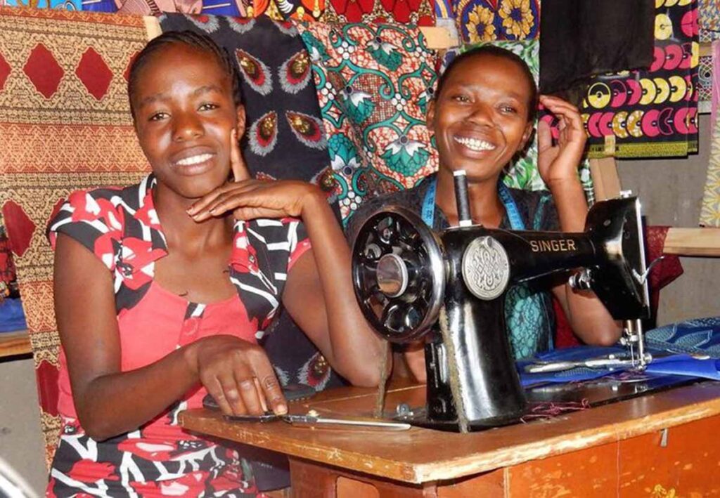 Two women smile while sitting at a sewing machine, surrounded by colorful patterned fabrics. Supported by the charity Tools For Self Reliance Zambia, both appear happy and engaged in their empowering work together.