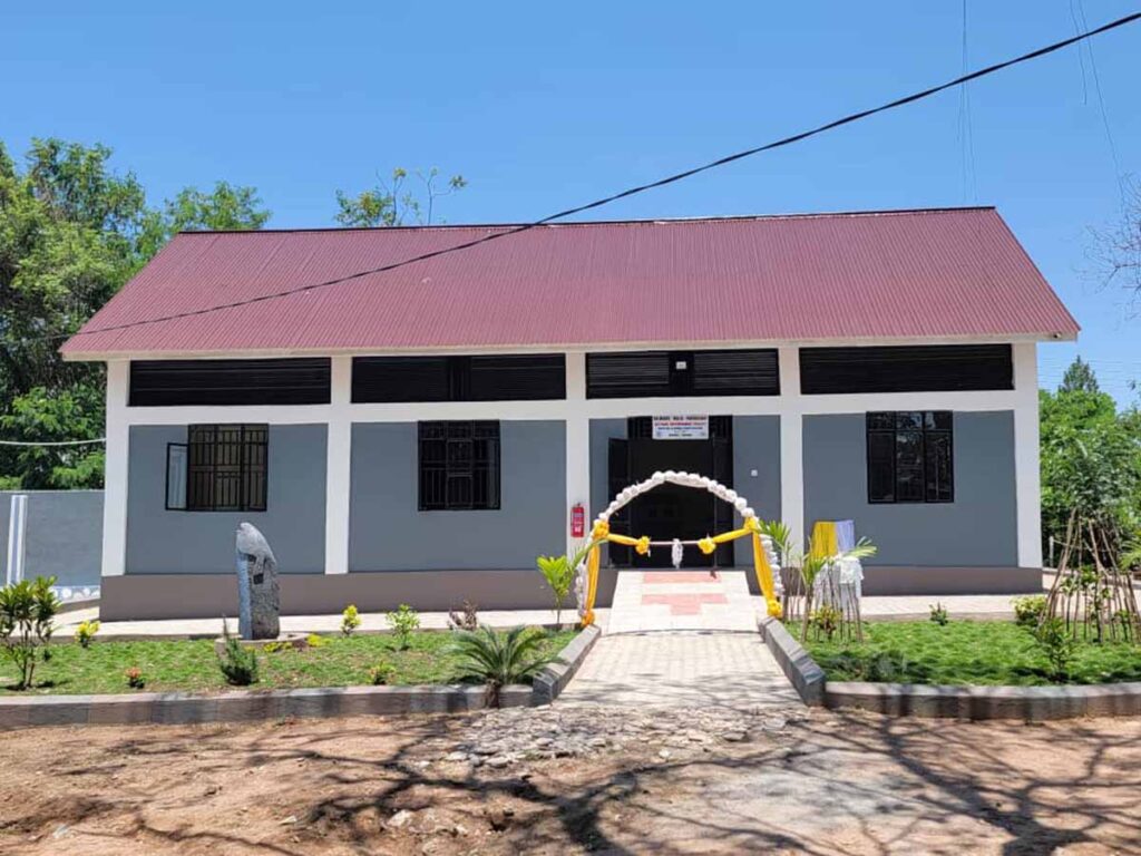 A small, single-story gray building with a red roof and black window frames. A decorated yellow and white arch marks the charity entrance, with a pathway leading up to it and a garden with plants in front.