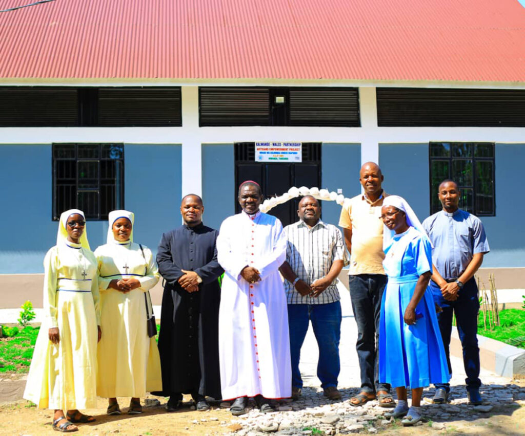 A group of eight people, including nuns, priests, and laymen, stand in front of a building with a red roof and blue walls. Smiling for the camera, they represent the Tools For Self Reliance charity in the sunlight.