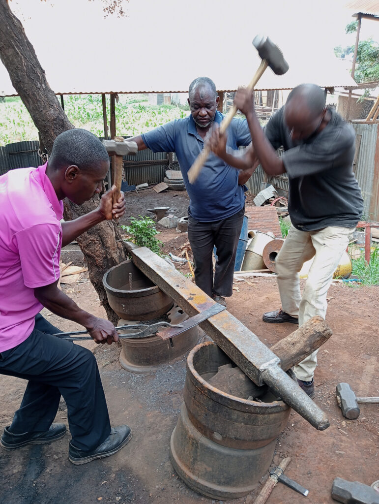 Three men in Zambia work together outdoors, forging metal on an improvised anvil. Using Tools For Self Reliance, one holds the metal steady, another hammers forcefully, while the third assists. Various tools and barrels are visible around them.