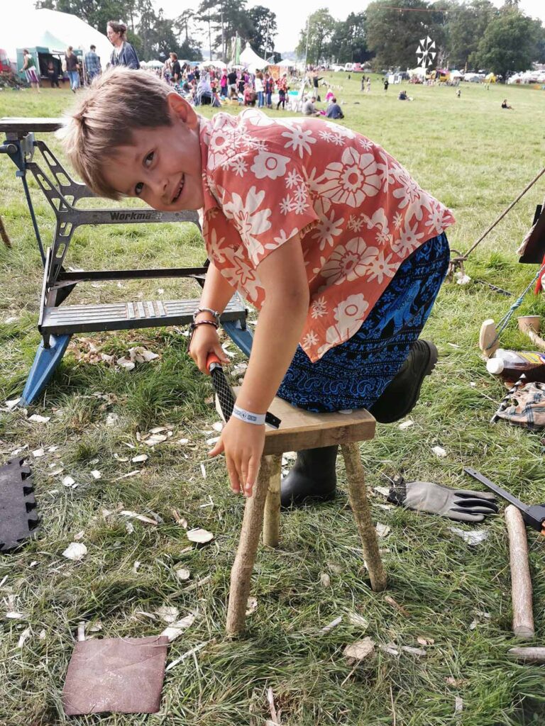 A smiling child in a floral shirt and patterned pants kneels on a small wooden stool in a grassy outdoor area, surrounded by Tools For Self Reliance woodworking tools and shavings, with tents and people in the background.