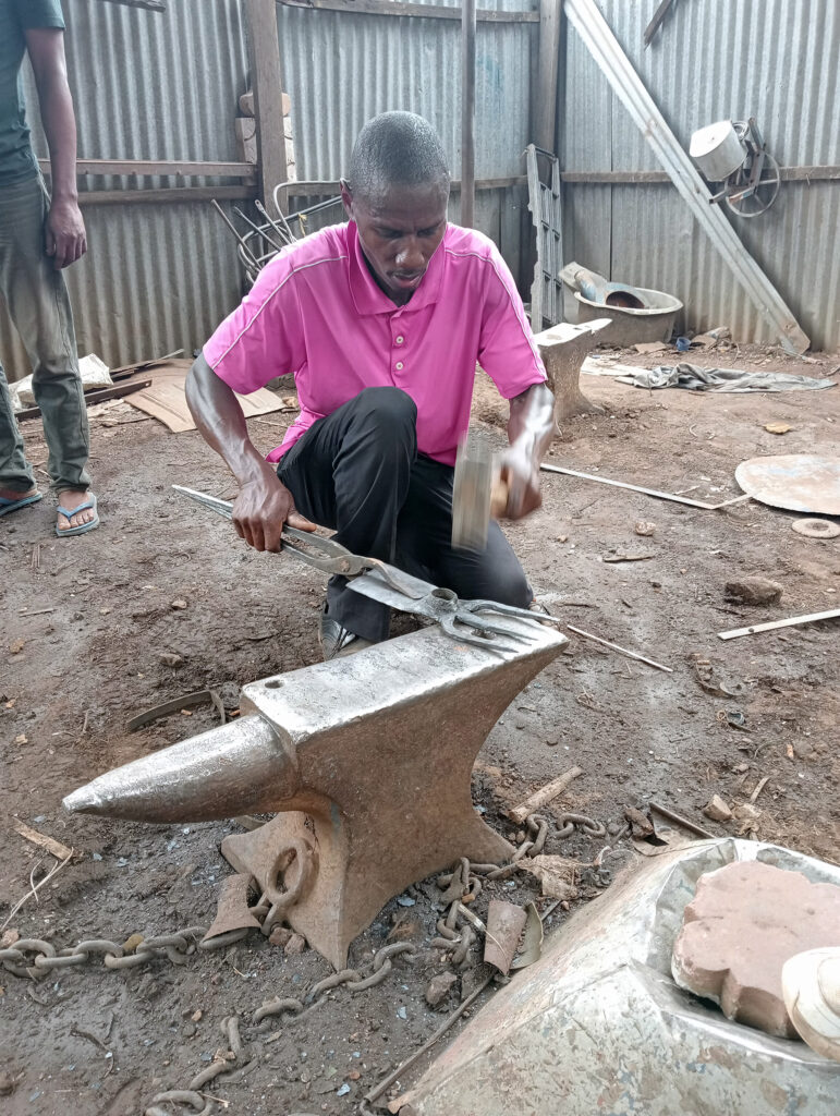 In Zambia, a man in a pink shirt uses a hammer and tongs to work on metal placed on an anvil inside a workshop with dirt floors and corrugated metal walls. Tools and other metal objects are scattered around in this corner of Africa.