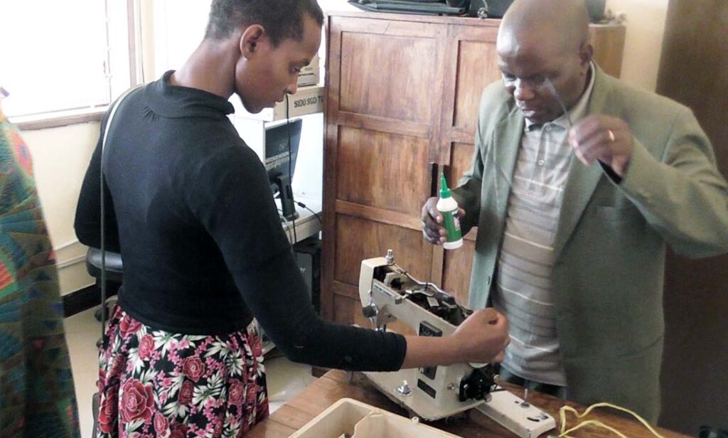 A woman threads a sewing machine as a man stands nearby holding a bottle, possibly of oil or glue, surrounded by wooden cabinets and a window—an everyday scene highlighting Tools For Self Reliance in Africa.