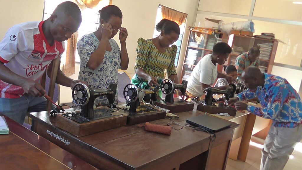 A group of people work together at a table using sewing machines and tools in a well-lit room with windows and shelves, showcasing the impact of Tools For Self Reliance in Zambia as they sew and repair equipment.