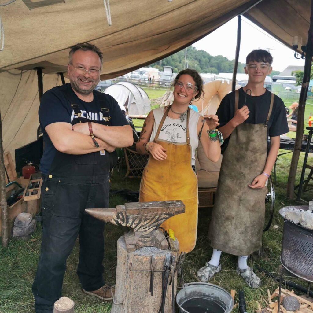 Three people stand in a tent at an outdoor event, smiling at the camera. Two wear aprons and have sooty faces, suggesting blacksmithing work for Tools For Self Reliance, a charity supporting skills development in Africa. An anvil and tools are visible nearby.