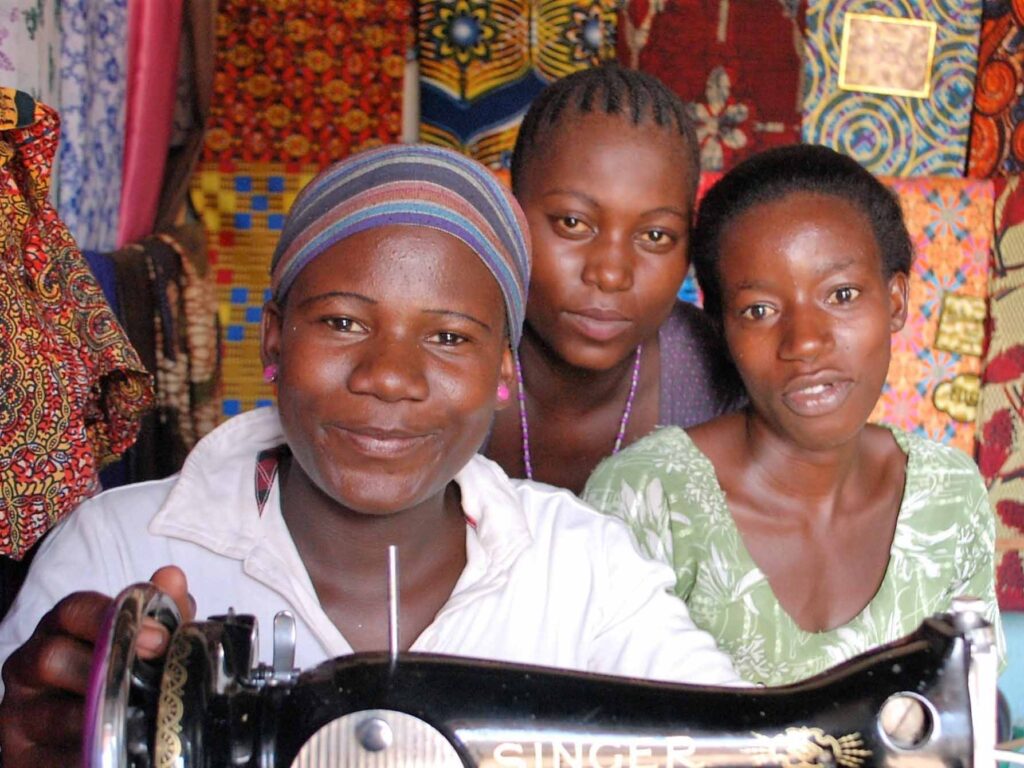 Three women smile together in front of colorful patterned fabric, with one holding a vintage Singer sewing machine—part of a Tools For Self Reliance charity project in Zambia.