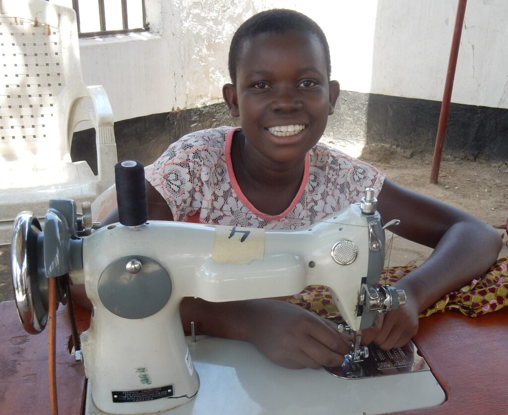 A smiling girl sits at a table using a sewing machine, with both hands on the fabric, indoors near a white plastic chair and wall—empowered by Tools For Self Reliance's support in Zambia.