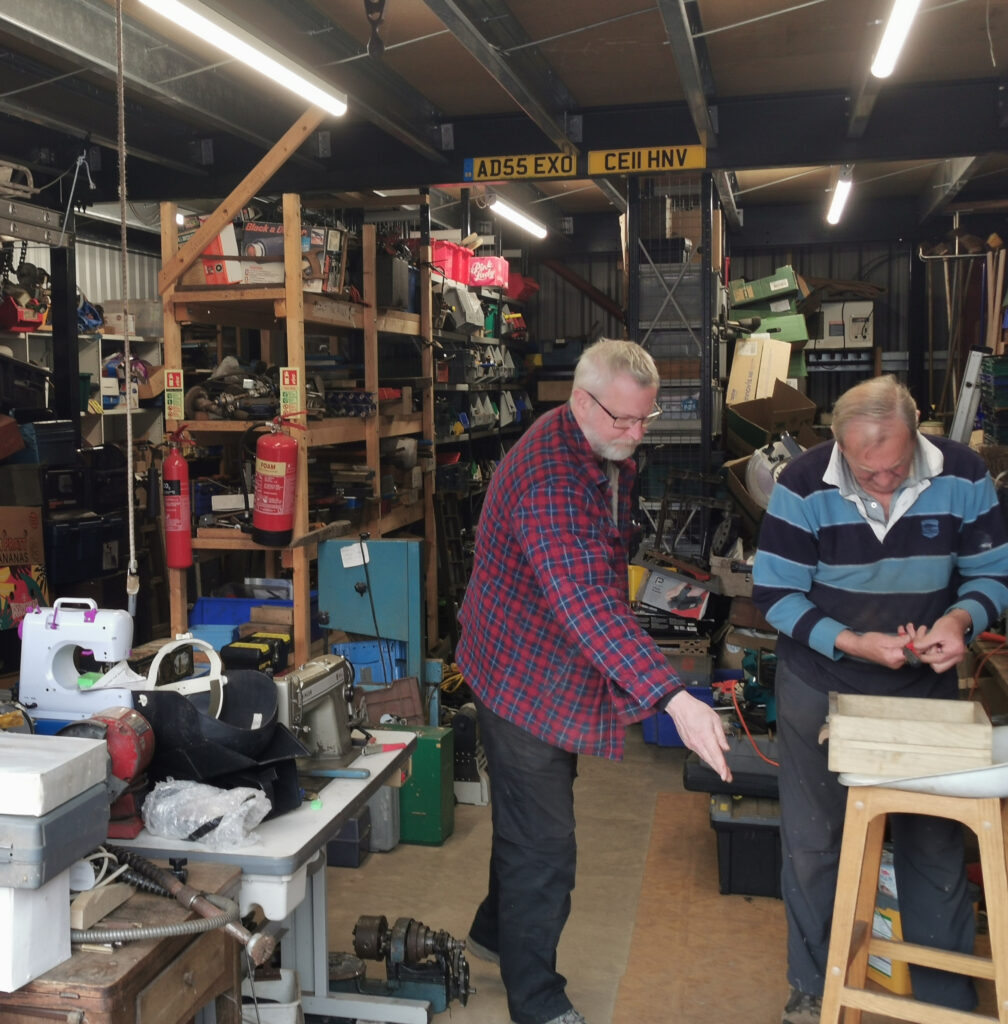 Two men work in a cluttered workshop filled with Tools for Self Reliance, shelves, and various equipment. One points towards something on a table, while the other works on a project, showcasing the charity's dedication to empowering others through tools.