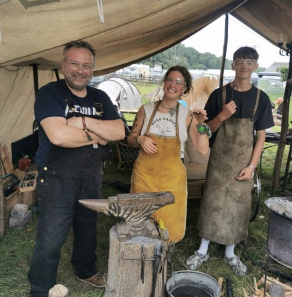 Three people stand smiling under a canvas tent at an outdoor event in Zambia, dressed in aprons and standing beside a blacksmith’s anvil and Tools For Self Reliance equipment, suggesting a demonstration or workshop.