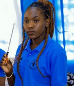 A young woman with braided hair wearing a blue shirt holds a tool with a black and orange handle, standing indoors with blue and white decorations in the background, representing a charity project in Zambia.