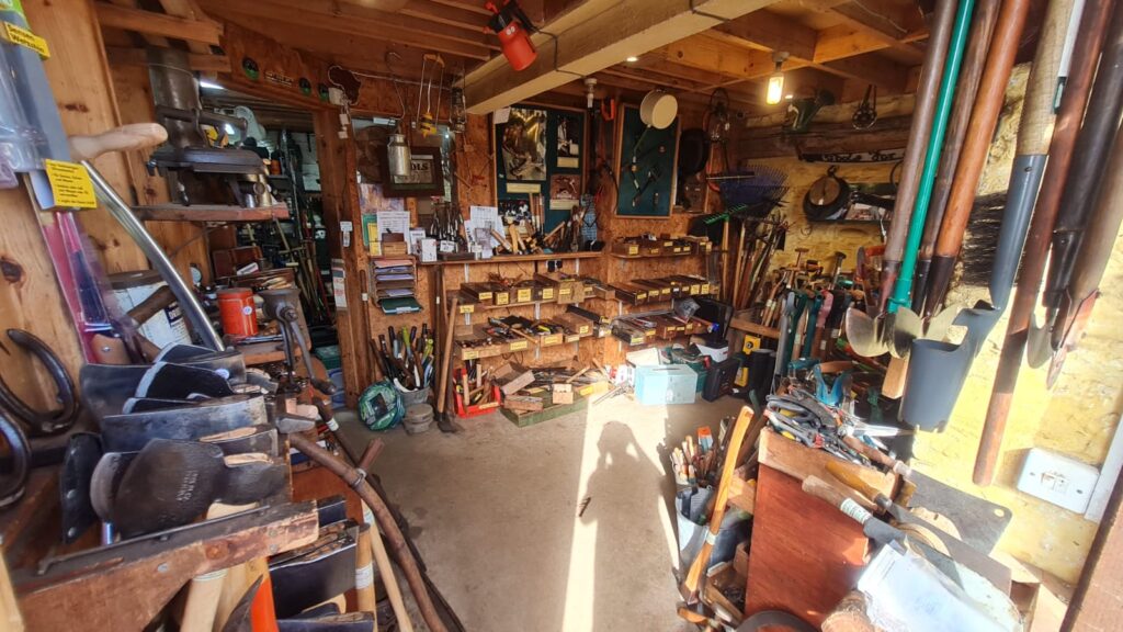 A well-organized workshop with shelves full of tools, boxes, and equipment. Shovels, rakes, and other gardening tools hang on the right wall, supporting a local charity’s projects in Africa as sunlight casts a shadow on the concrete floor.