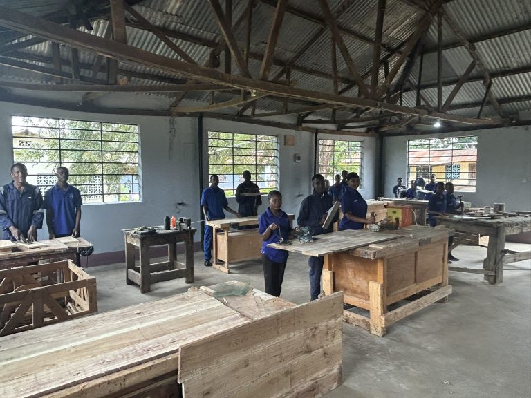 A group of students in blue uniforms works at wooden tables in a spacious, well-lit workshop with large windows and exposed metal beams. Various woodworking tools and materials are visible on the tables.