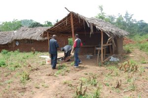 Three people stand near a simple, partially constructed hut with a thatched roof in a rural area. Another similar building is visible in the background amid green vegetation and earth ground.