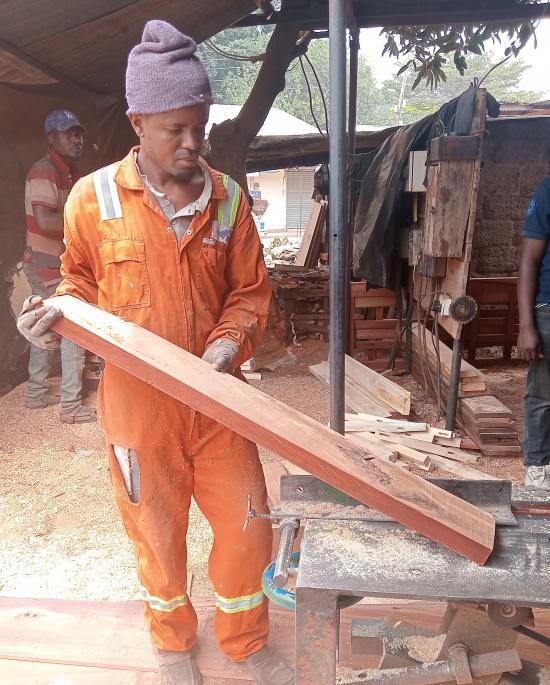 A man in an orange work overall and grey beanie is holding a wooden plank by a saw in a workshop, with sawdust on the floor and other workers and wooden materials in the background.