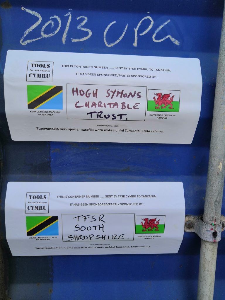 Two white labels on a blue surface show Tanzanian and Welsh flags, charity logos, and text about donations from "Hugh Symons Charitable Trust" and "TFSR South Shropshire" to Tanzania. "2013 UPC" is written above in chalk.