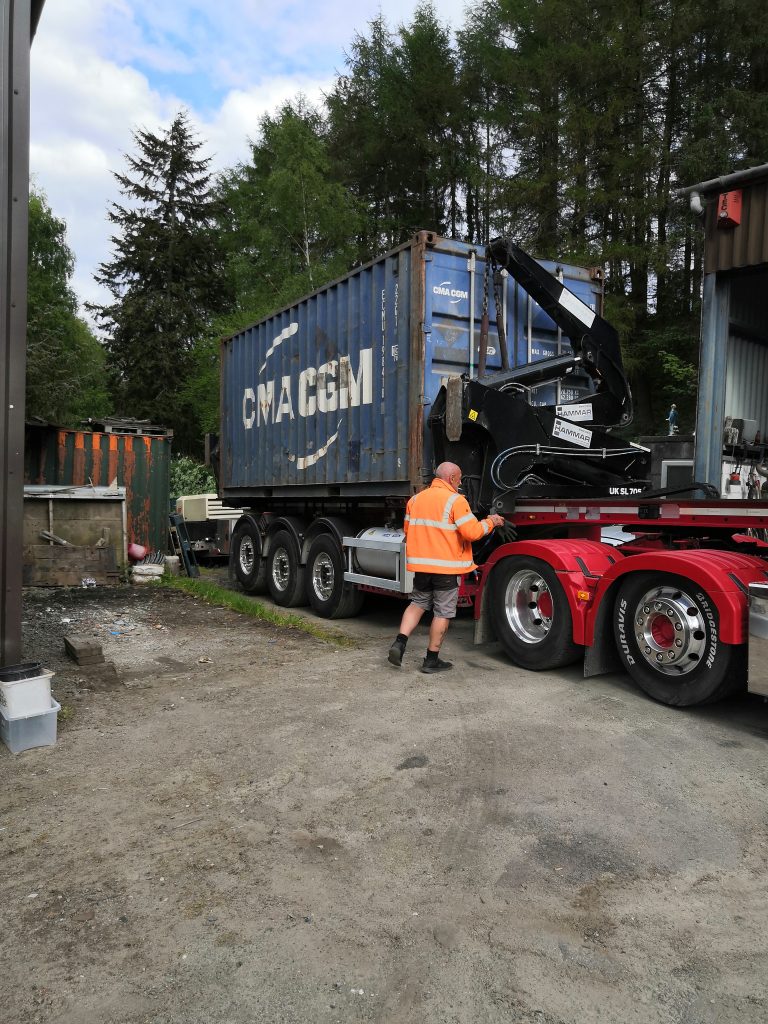A person in an orange high-visibility vest stands next to a red lorry with a large blue CMA CGM shipping container in a yard surrounded by trees and various equipment.