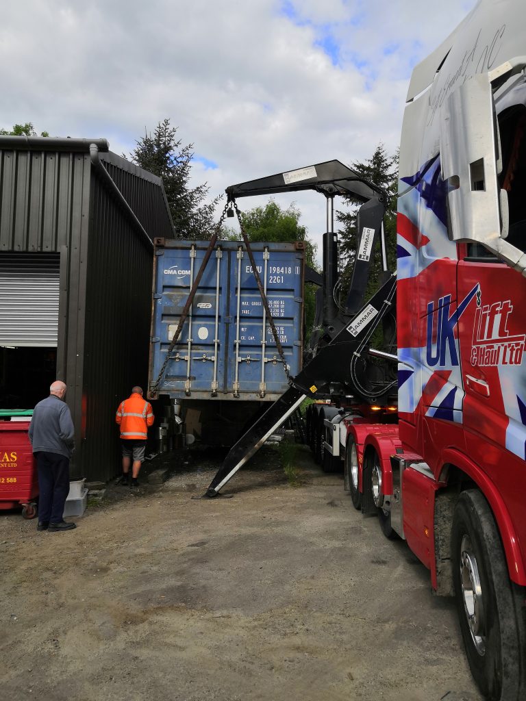 A blue shipping container is being unloaded from a lorry by a hydraulic lift next to a dark metal building. Two people, one in an orange jacket, stand nearby, observing the process.