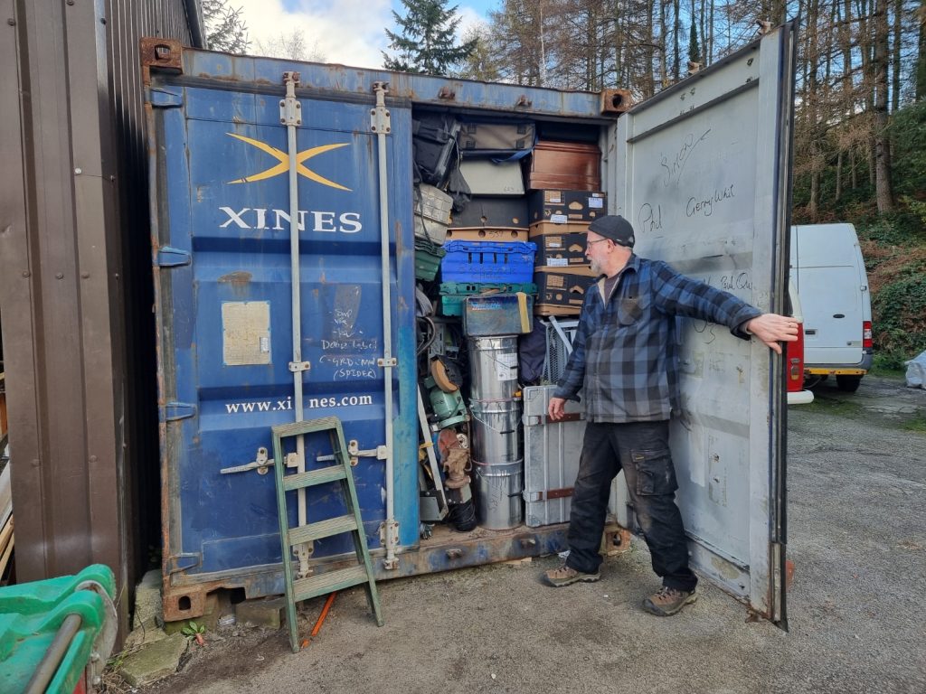 A man in a cap and chequered jacket stands by the open door of a blue shipping container packed full of various boxes, tools, and equipment, with trees and a white van in the background.