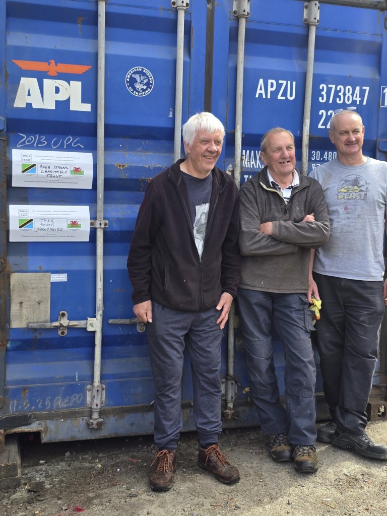 Three older men stand smiling in front of a large blue shipping container with "APL" and codes visible. Two paper signs are attached to the container door. The men wear casual clothing and appear relaxed and happy.