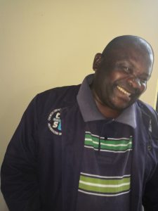 A man wearing a navy jacket and a striped green and grey shirt smiles warmly and tilts his head, standing in front of a plain light-coloured wall.