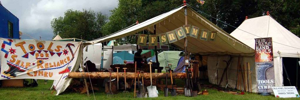 A festival marquee labelled "TOOLS CYMRU" displays various hand tools on a wooden log. Colourful signs read "Tools for Self Reliance Cymru" and "Fair Trade Hand Tools." Other marquees and trees are visible in the background.