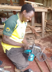 A man kneels on the workshop floor, smiling as he holds a circular saw. Wood and tools are visible around him, suggesting he is engaged in joinery work.