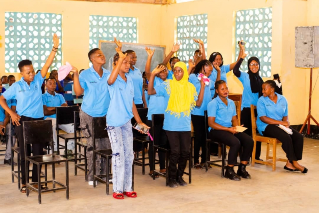 A group of young people in blue shirts stand and sit in a classroom, with several pupils raising their hands.
