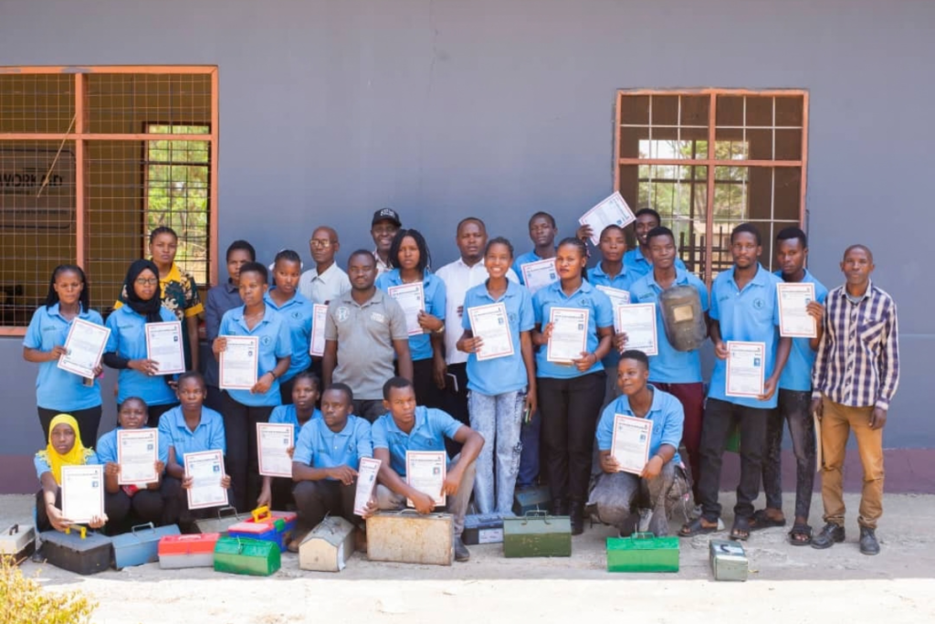 A large group of students in light blue shirts stand in front of a building holding certificates, with several metal toolboxes on the ground in front of them.